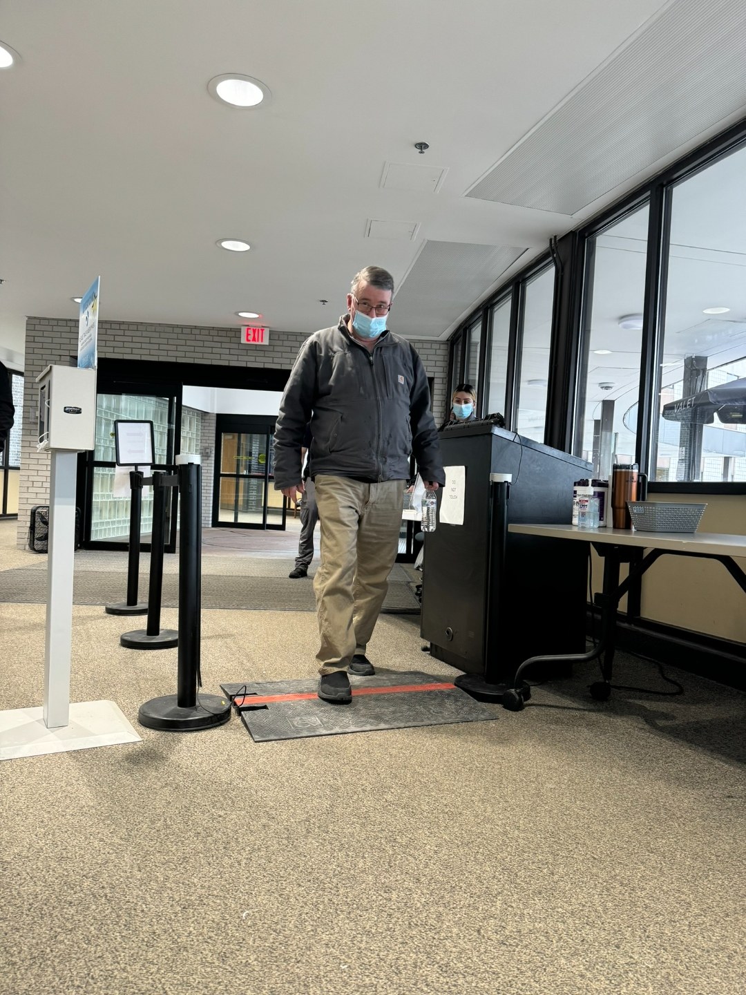 BallistiSCAN at the public main entrance of Upstate University Hospital in Syracuse, New York — a visitor walking through the passive scan mat between two stanchion-mounted sensors with the security desk visible to the side.